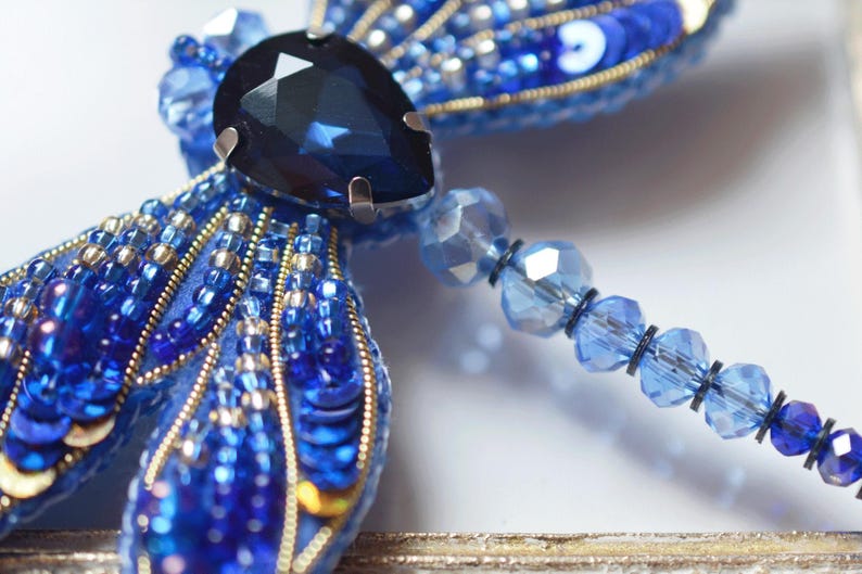 May include: A handcrafted, blue dragonfly brooch. The wings are embellished with sequins and beads in shades of blue and gold. A large, faceted, dark blue gem is at the center. A beaded strand extends from the dragonfly's body.