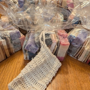 May include: Bundles of colorful handmade soaps wrapped in clear cellophane, tied with twine, and a beige woven soap saver. The soaps are various colors, including purple, pink, and beige. The soap saver is made of a loosely woven material.