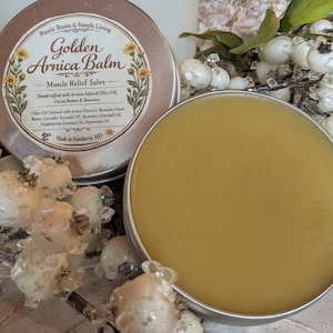 May include: A close-up of two open, round metal tins of Golden Arnica Balm. The balm is a yellow-gold color. The lid of one tin is visible, with text that reads "Golden Arnica Balm - Muscle Relief Salve". The product is handcrafted with Arnica-infused olive oil.