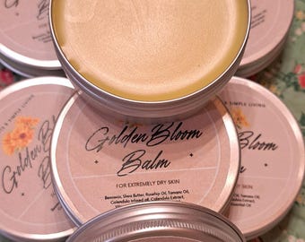 May include: Several round, silver tins of Golden Bloom Balm for extremely dry skin. The balm has a pale yellow hue and is labelled with the product name and ingredients. The background features a floral pattern.