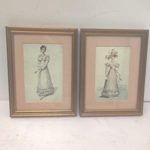 May include: Two framed prints featuring illustrations of women in period clothing. The prints are in gold-colored frames with a pink matting. The illustrations depict women in long dresses and bonnets, with delicate details and soft colors.