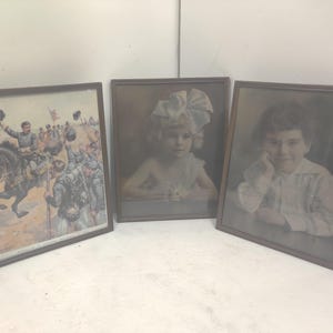 May include: Three framed vintage prints. One depicts a military scene with a man on horseback, another a young girl with a large bow, and the third a portrait of a child. All are in brown wooden frames.