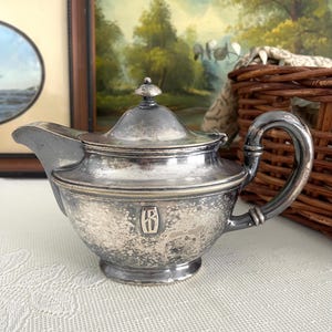May include: A tarnished silver-toned metal teapot featuring a curved handle, a short spout, and a removable lid with a small finial. The body of the teapot displays a prominent embossed monogram featuring the letters IB enclosed within a rectangular frame.