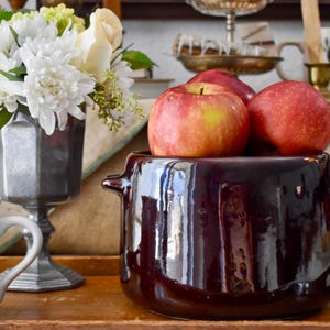 May include: A still life arrangement featuring a dark brown ceramic pot filled with red apples. A silver vase holds white flowers, and a floral-decorated mug sits nearby. The scene is set on a wooden tray.