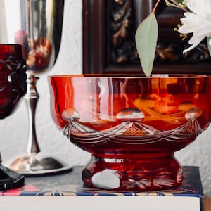 Puede incluir: Una colección de cristalería roja rubí vintage. La imagen presenta una copa, un cuenco y un vaso alto con pie. El cristal rojo rubí tiene intrincados patrones tallados. El cuenco está sobre una pila de libros, y un jarrón de flores blancas está en el fondo.