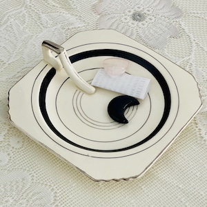 May include: A cream-colored, octagonal plate with black concentric circles and a silver rim. The plate holds a white handle with a silver accent, a pink crystal, a small piece of paper, and a black crescent moon-shaped object.