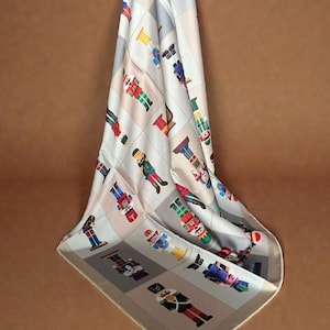 May include: A light-colored scarf featuring a repeating grid pattern of various nutcracker figures in red, blue, green, and gold uniforms. The nutcrackers are depicted in different poses and colors, arranged in a structured layout across the fabric.