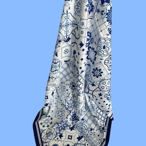 May include: A white scarf with a blue geometric tile pattern. The scarf has a dark blue border and features various floral and geometric designs. The scarf is made of a soft, flowing fabric.