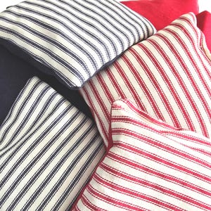 May include: Close-up of several throw pillows in various colors and patterns. Some pillows are navy blue, others are red, and some have white and navy blue or red stripes. The pillows are arranged in a casual, overlapping manner.