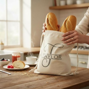 May include: A beige canvas drawstring bag with the name "TAYLOR" printed on it, holding two loaves of bread. The bag is on a wooden surface, with a plate of food and a cup of coffee in the background.