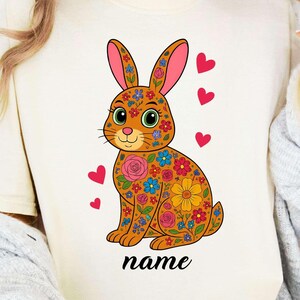 Puede incluir: Camiseta color crema con un conejito con estampado floral, orejas rosas y detalles de corazón. El conejito está adornado con flores coloridas y la palabra "name" está escrita en cursiva debajo.