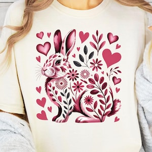 Puede incluir: Camiseta color crema con un diseño floral de conejo rosa y blanco, corazones y detalles florales. El diseño incluye una ilustración detallada de un conejo rodeado de flores y corazones, ideal para Pascua o primavera.