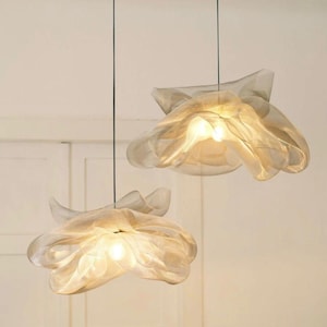 May include: Two pendant lights with a soft, flowing design. The lampshades are a translucent, off-white material, resembling flower petals. Each light has a single bulb, emitting a warm glow. The lights hang from black cords against a neutral background.