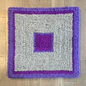 Puede incluir: Una alfombra cuadrada con una construcción de bucles texturizados. El diseño consiste en un cuadrado central morado, rodeado por un borde ancho de bucles gris claro, encerrado por un borde morado fino y un marco exterior morado.