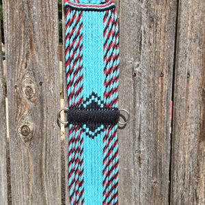 May include: A woven horse cinch featuring a bright turquoise base with vertical stripes of black and red twisted cord. The centre displays a black diamond pattern, and the ends are finished with silver-toned metal buckles and side rings.