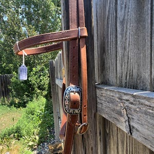 May include: A brown leather horse bridle featuring white stitching and a silver-toned metal buckle with an engraved scrollwork pattern. The bridle includes a browband and cheek pieces with adjustable holes, hanging vertically from a wooden post.