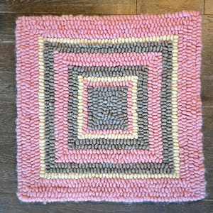 Könnte beinhalten: Quadratischer rosa Teppich mit geometrischem Design. Der Teppich hat ein zentrales Quadrat in Grau, umgeben von einem cremefarbenen Rand, dann einem grauen Rand und schließlich einem rosa Rand. Der Teppich besteht aus einem strukturierten Material.
