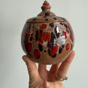May include: A ceramic jar with a lid, displaying a brown base with red and black speckled patterns. The lid has a small, pointed finial. The jar is held in a hand, highlighting its size and design.