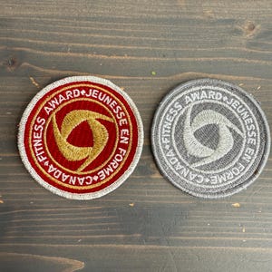 Peut inclure: Deux écussons ronds brodés. L'un est rouge avec des accents dorés et blancs, l'autre est gris avec des accents blancs. Les deux portent l'inscription "Fitness Award Jeunesse en Forme Canada" et un motif stylisé au centre.