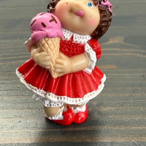 May include: A small figurine of a doll with brown curly hair and a pink bow, wearing a red dress with white lace trim. The doll has blue eyes and rosy cheeks, and is holding a pink ice cream cone with brown sprinkles in its hands.