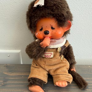 May include: A vintage Monchhichi doll with brown fur, wearing corduroy overalls. The doll has a pink bib, a small tuft of hair tied with a pink ribbon, and the number "007" printed on the overalls. The doll is sitting on a wooden surface.