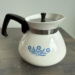 May include: White vintage coffee pot with a black handle and lid. The pot has a blue floral design and a silver band. The coffee pot is on a dark wooden surface.