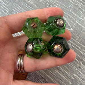 May include: Four green glass cabinet knobs with a faceted, hexagonal design. Each knob has a metal screw and a metal cap. The knobs are a vibrant shade of green and appear to be vintage or antique.