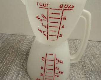 May include: White measuring cup with red markings for ounces and cups. The cup has a handle and a spout for easy pouring. The markings include measurements like 1 cup and 8 oz.
