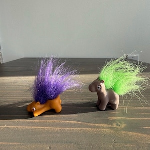 May include: Two troll dolls with spiky, colourful hair. One is orange with purple hair, the other is brown with green hair. They have small, round bodies and are posed on a wooden surface.