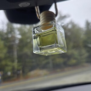 Puede incluir: Un ambientador de coche transparente, de forma cuadrada, con una tapa de madera y una cuerda beige para colgarlo. El l&iacute;quido del interior es de color amarillo verdoso claro. El ambientador cuelga del espejo retrovisor de un coche.
