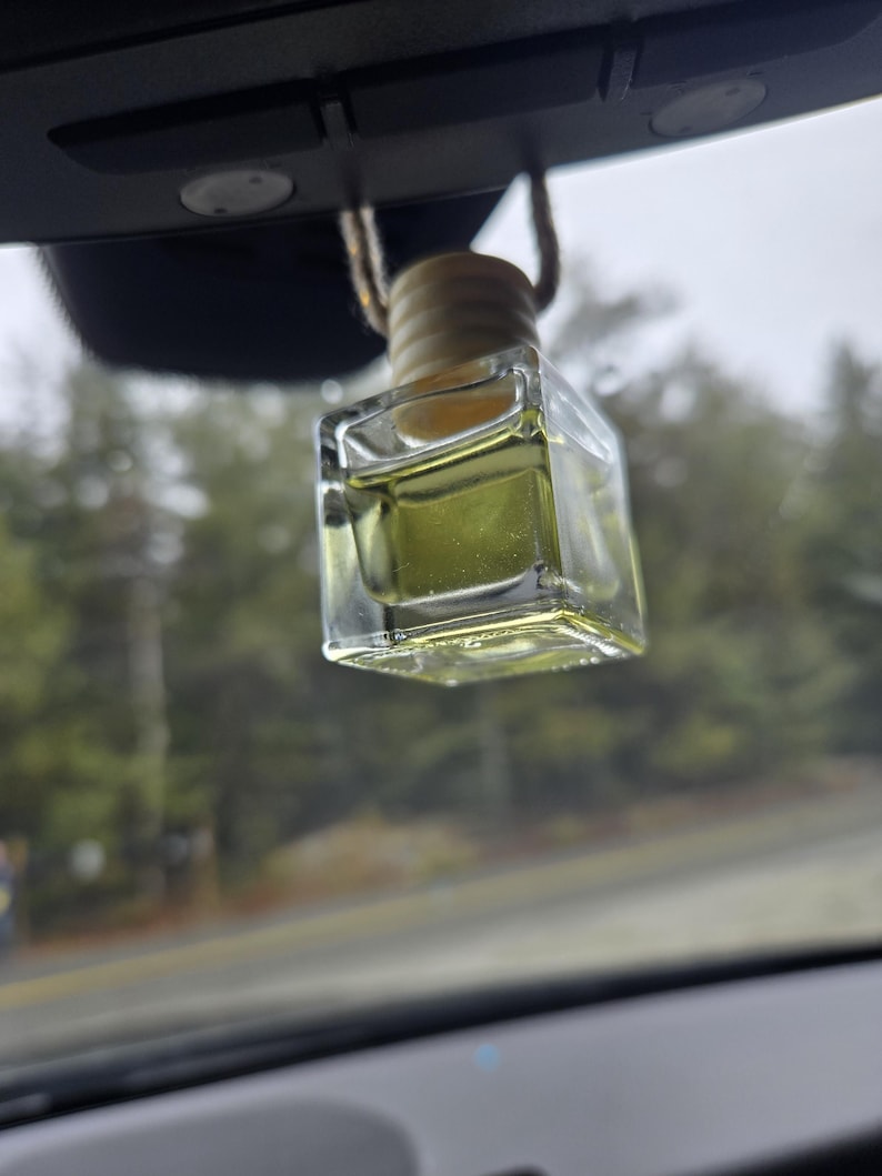 Puede incluir: Un ambientador de coche transparente, de forma cuadrada, con una tapa de madera y una cuerda beige para colgarlo. El l&iacute;quido del interior es de color amarillo verdoso claro. El ambientador cuelga del espejo retrovisor de un coche.