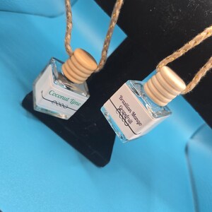 Puede incluir: Dos peque&ntilde;os ambientadores para coche de cristal transparente con tapones de madera y cordones de cuerda. Uno dice "Coconut Lime", el otro "Brazilian Mango Grapefruit". Los ambientadores cuelgan sobre un fondo negro en una superficie azul.