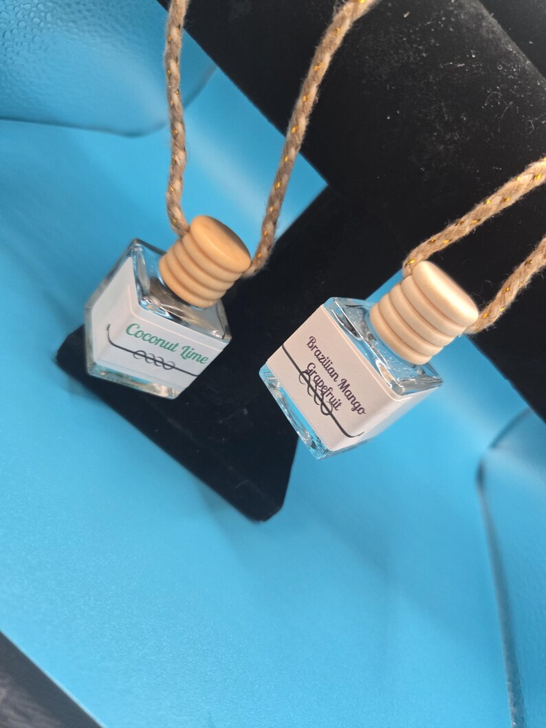Puede incluir: Dos peque&ntilde;os ambientadores para coche de cristal transparente con tapones de madera y cordones de cuerda. Uno dice "Coconut Lime", el otro "Brazilian Mango Grapefruit". Los ambientadores cuelgan sobre un fondo negro en una superficie azul.