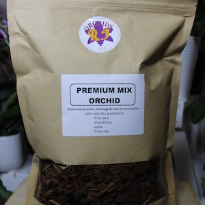 May include: A brown paper bag with a clear window showing a mix of orchid potting medium. The bag has a label that reads "Premium Mix Orchid" and lists the ingredients: pine bark, coco chips, leca, and charcoal. The label also states that the mix is 100% natural and organic.