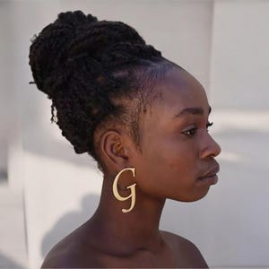 Könnte beinhalten: Eine Person mit dunkelbraunen Haaren in einem Dutt trägt einen goldenen Ohrring in Form des Buchstabens G.