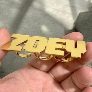 May include: Gold-toned adjustable ring set with the name "ZOEY" spelled out in block letters.