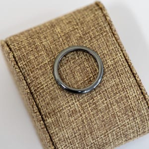 May include: A dark gray ring with a smooth, polished surface. The ring is circular and sits on a textured, brown fabric surface. The ring's color is a deep, metallic gray, and it appears to be made of a solid material.