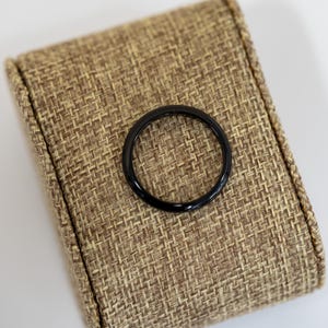 以下が含まれることがあります： 光沢のある黒いリングが、質感のあるベージュ色の表面に置かれています。リングは円形で、しっかりとした素材でできているようです。背景はニュートラルな色で、リングの暗い色合いを引き立てています。