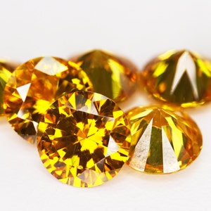 May include: A group of seven bright yellow gemstones, likely diamonds, are arranged on a white surface. The gemstones are faceted and reflect light, creating a sparkling effect.