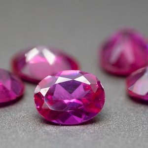 Puede incluir: Cinco piedras preciosas ovaladas facetadas en un tono rosa vibrante. Las piedras preciosas están dispuestas sobre una superficie gris.
