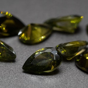 May include: A collection of pear-shaped green gemstones, some with a slight golden hue, displayed on a dark gray surface.