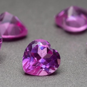 Op de afbeelding: Close-up van verschillende hartvormige, gefacetteerde edelstenen in roze en paarse tinten. De edelstenen liggen op een grijze ondergrond, met lichtreflectie op hun geslepen oppervlakken. De edelstenen zijn voor sieraden.