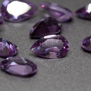 May include: A group of purple pear-shaped gemstones on a gray surface.