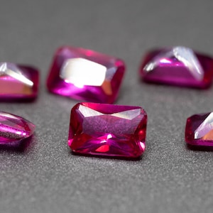May include: Six pink rectangular gemstones with a faceted cut. The gemstones are arranged on a gray surface.