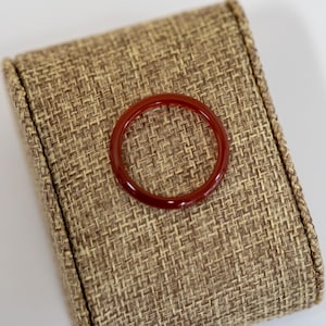 May include: A polished, translucent red ring. The ring is circular and sits on a textured, tan fabric surface. The ring's color is a deep, rich red, and it appears to be made of a smooth, glossy material. The ring is a piece of jewelry.
