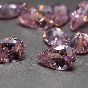 May include: A group of pear-shaped pink gemstones, some with a more rounded shape, are scattered on a gray surface. The gemstones are faceted and reflect light.