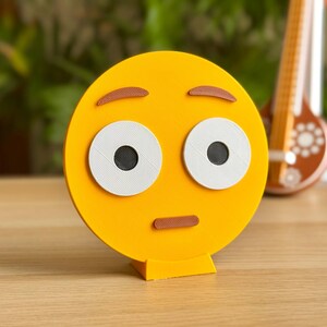 May include: A yellow 3D printed emoji with a surprised expression. The emoji has large white eyes with black pupils, brown eyebrows, and a brown mouth. The emoji is standing on a small yellow base.