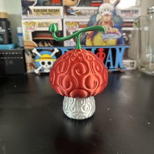 May include: A red and white 3D printed figurine of a mushroom with a green stem. The mushroom has a textured surface and a white base.