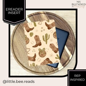 May include: An e-reader insert with a beige background featuring brown cowboy boots, cacti, and other western-themed illustrations. The insert is placed on a wooden tray with a dark blue e-reader peeking out from behind it. Text on the insert reads "EREADER INSERT".