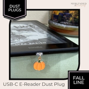 May include: A small orange pumpkin charm attached to a clear plastic USB-C dust plug, inserted into the charging port of a black e-reader. The pumpkin features a green stem detail and is suspended from a gold-toned metal jump ring.